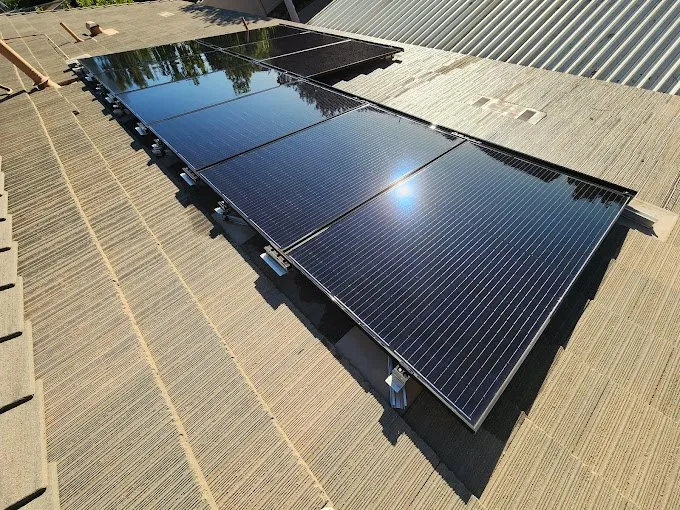 Rows of solar panels are installed on a corrugated rooftop under bright sunlight, reflecting the sky and some nearby trees.