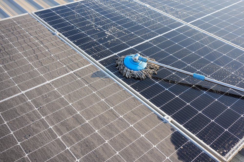 A rotating brush attached to a long pole is cleaning dust and dirt from solar panels on a rooftop, with a clear contrast between cleaned and dirty sections.