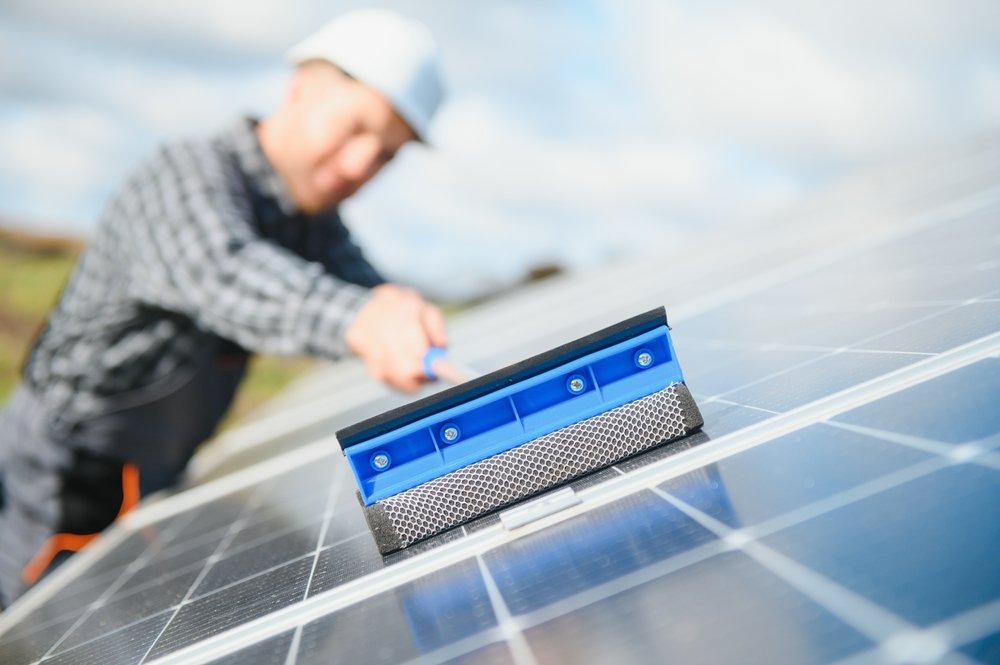 A person wearing a hard hat uses a squeegee to clean the surface of solar panels outdoors on a sunny day, with the focus on the panels and cleaning tool.