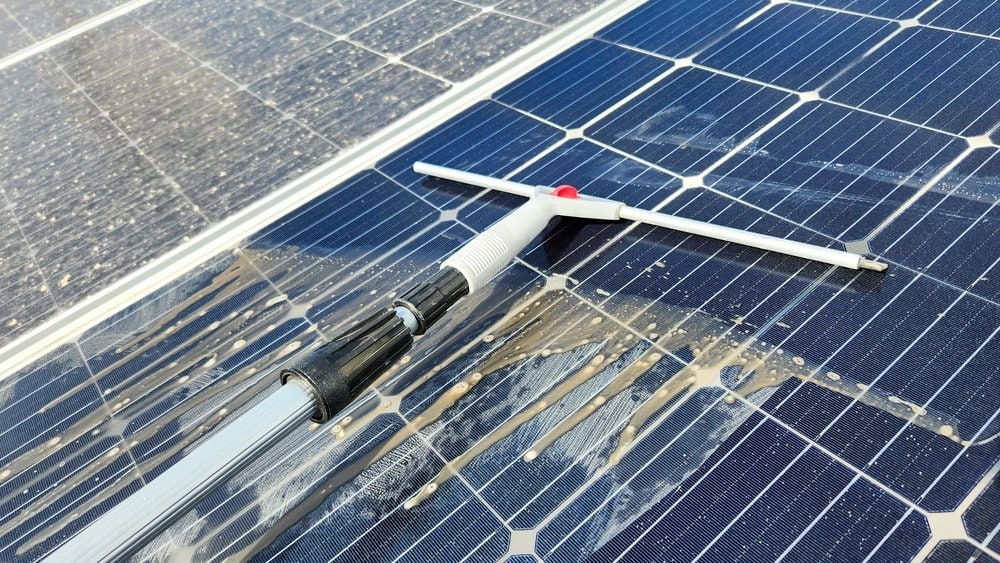A close-up of a squeegee cleaning dirt and dust from the surface of solar panels, revealing the shiny blue cells beneath. The cleaning tool moves across the panels, leaving a clear, streak-free area.