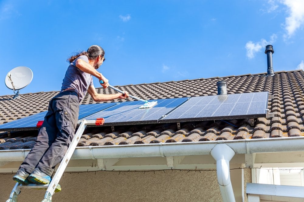 A person standing on a ladder cleans solar panels installed on a tiled house roof under a bright blue sky.