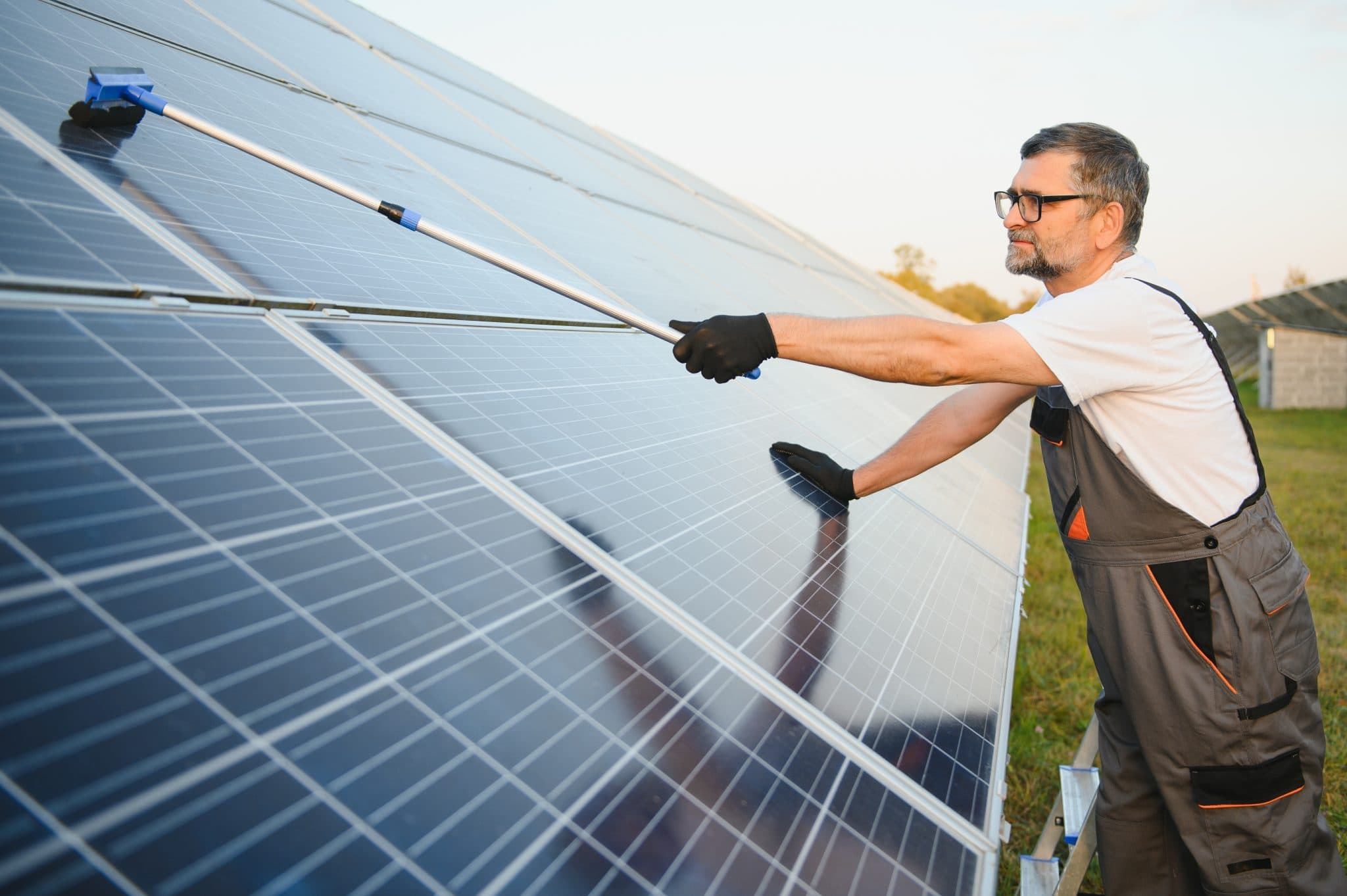 A man wearing gloves and overalls cleans a large outdoor solar panel with a long-handled brush under sunlight.