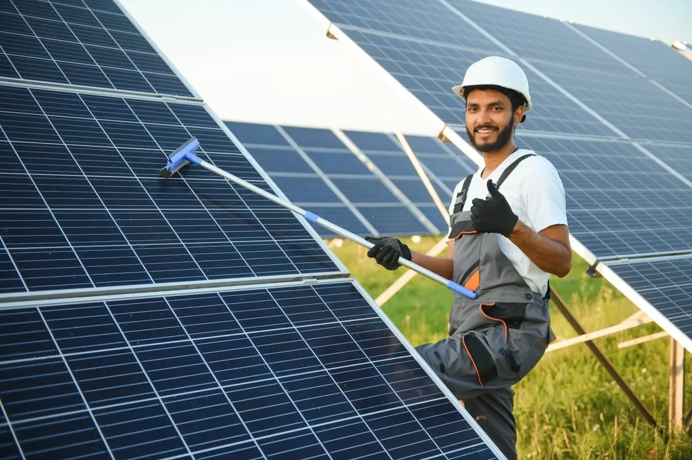 A man in work overalls and a white helmet cleans solar panels with a squeegee outdoors, smiling and giving a thumbs-up gesture on a sunny day.