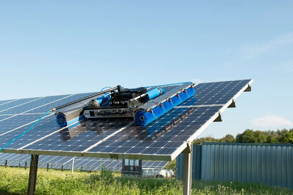 A robotic cleaning device with blue brushes is moving over large solar panels outdoors on a sunny day, cleaning the surface. Green grass and other solar panels are visible in the background.
