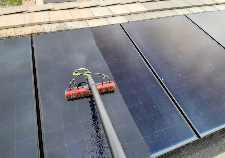 A large brush on a long handle is cleaning rooftop solar panels, with a clear difference between the washed and unwashed areas.