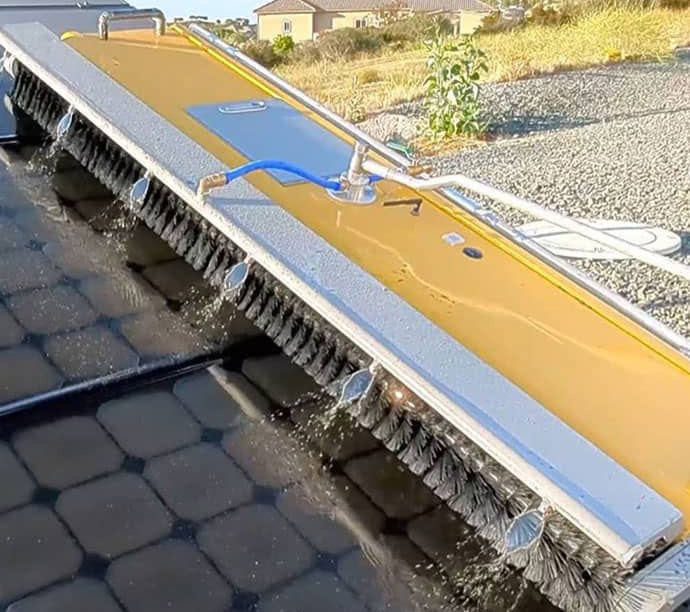 A large mechanical brush with water jets cleans solar panels outdoors on a sunny day, with gravel and vegetation visible in the background.