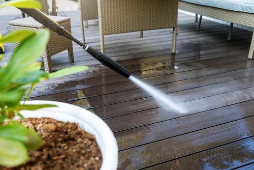 A person uses a pressure washer to clean a wooden deck outdoors, with patio furniture and a potted plant visible nearby.