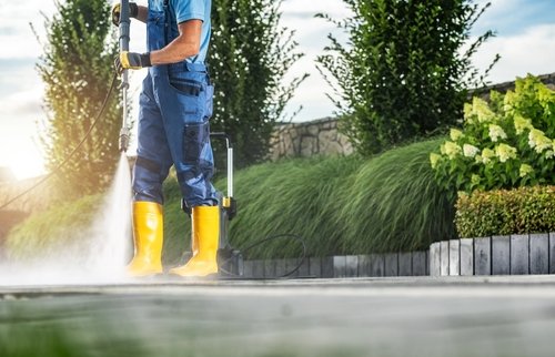 A person wearing blue overalls and yellow rubber boots uses a pressure washer to clean an outdoor pavement, with green bushes and trees in the background.
