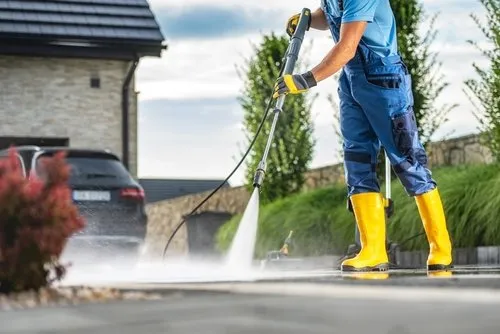 A person in blue overalls and yellow boots uses a pressure washer to clean an outdoor concrete surface near a house and a parked car, with green shrubs and trees in the background.