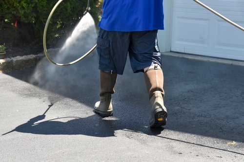 A person wearing tall boots, shorts, and a blue shirt power washes a driveway, spraying water onto the pavement near a garage door.