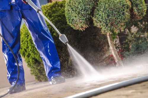 A person in blue waterproof overalls uses a power washer to clean an outdoor patio or walkway, with bushes and trimmed shrubs visible in the background.