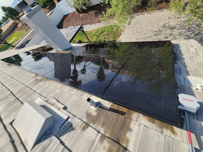 A rooftop with several black solar panels installed, reflecting the sky and trees, alongside a chimney and rooftop equipment. Gravel landscaping and greenery are visible in the background.