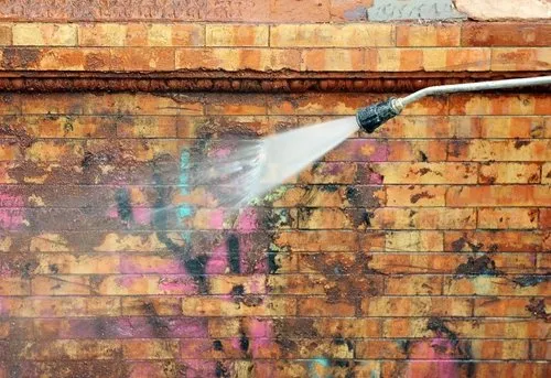 A power washer sprays water onto a brick wall covered in graffiti, removing paint and revealing the original brick surface underneath.