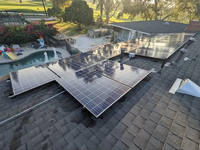 Rooftop solar panels installed on a shingled roof, with a backyard pool, patio area, and trees visible in the background on a sunny day.