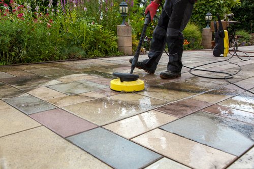A person is using a yellow power washer to clean a colorful tiled patio. Half of the tiles are clean while the other half are still dirty. Lush green garden plants are visible in the background.