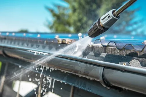 A pressure washer sprays water to clean the edge of a metal roof and gutter system on a sunny day, with trees visible in the blurred background.