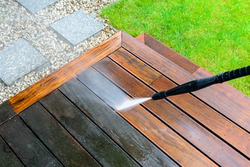 A person uses a pressure washer to clean a wooden deck, revealing a lighter, cleaner surface compared to the dirtier, darker wood beside it. Green grass and stone pavers are visible in the background.