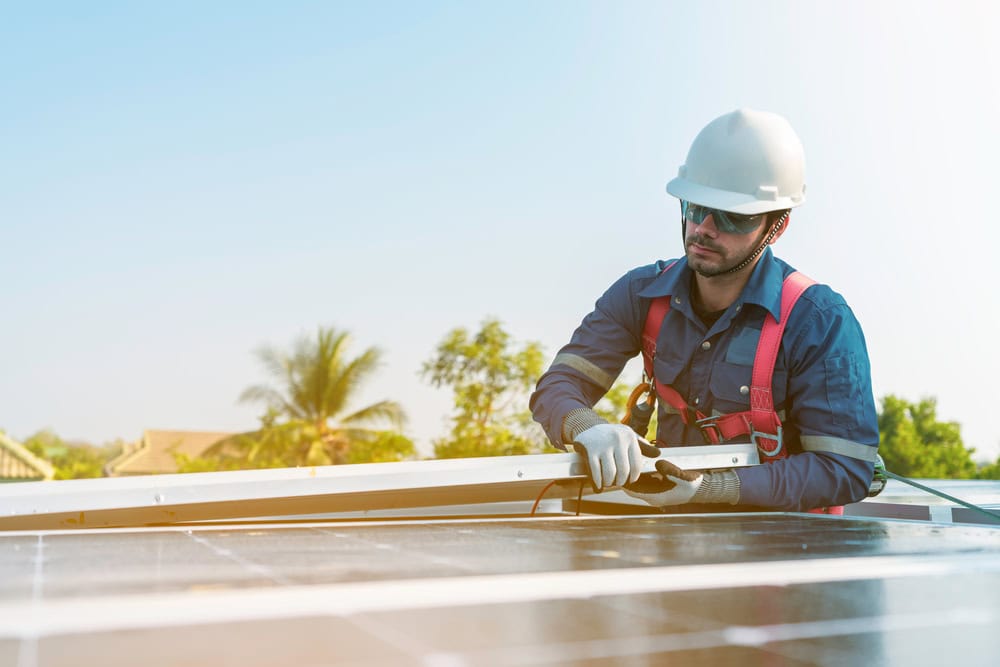 A worker in safety gear and a helmet installs a solar panel on a rooftop under a clear sky, with trees and buildings visible in the background.