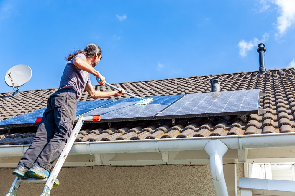 A person standing on a ladder cleans solar panels on a tiled house roof under a clear blue sky. A satellite dish and roof vents are also visible.