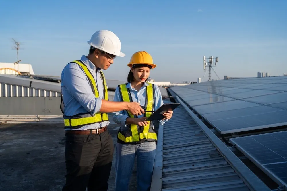 Two engineers wearing safety helmets and reflective vests stand on a rooftop beside solar panels, reviewing information on a tablet together under a clear blue sky.