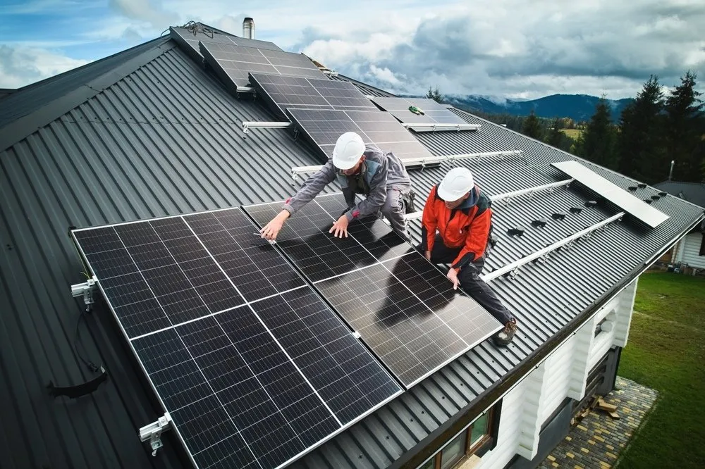 Two workers wearing safety helmets install solar panels on the black metal roof of a house, surrounded by trees and mountains under a cloudy sky.