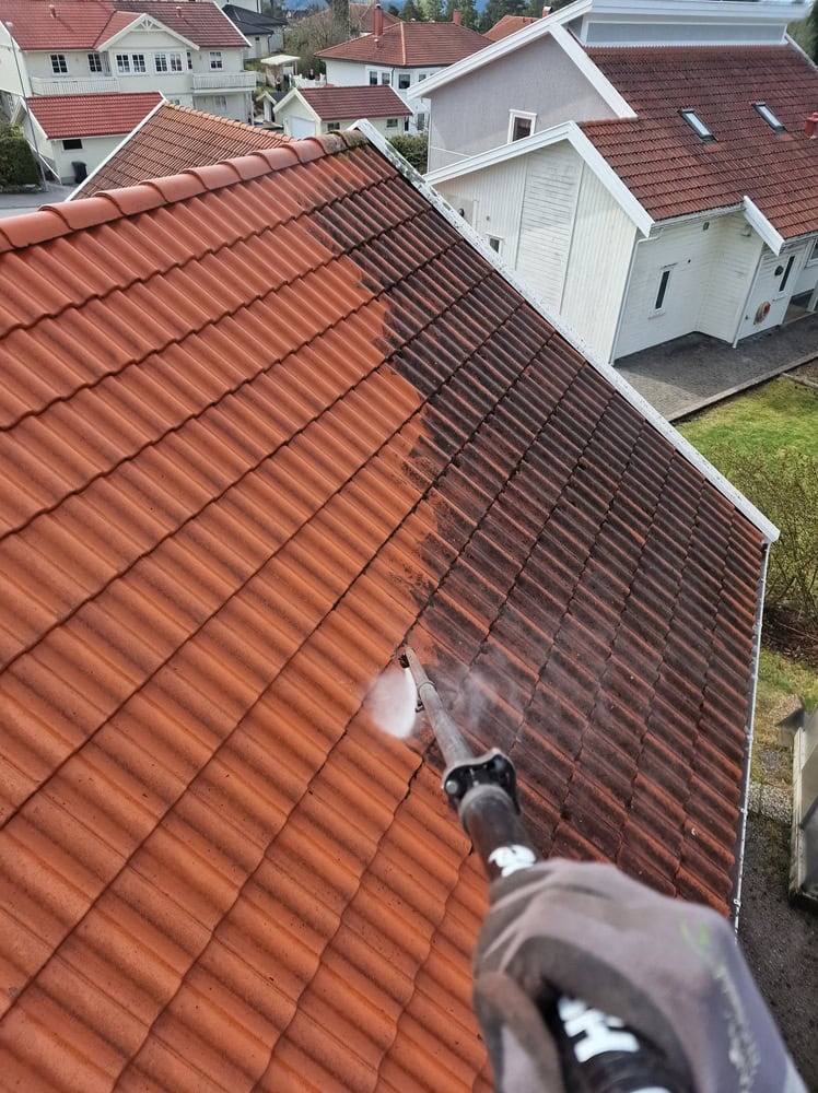 A person uses a power washer to clean a clay tile roof, with a clear contrast between the cleaned orange tiles and the still-dirty, darkened tiles. Several houses are visible in the background.