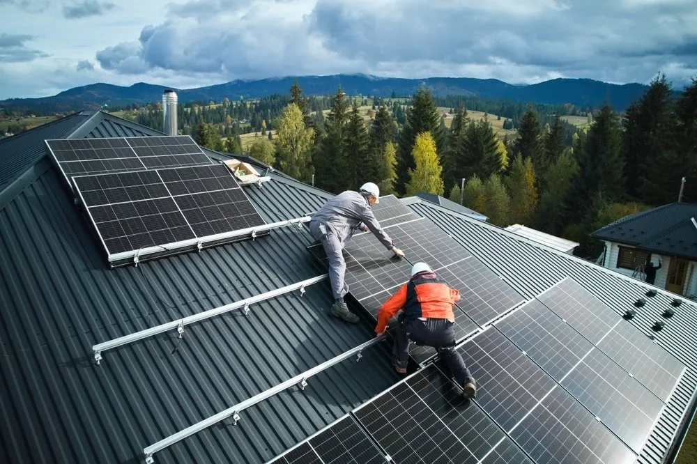 Two workers in safety gear install solar panels on a sloped metal roof, surrounded by trees and mountains under a cloudy sky in a rural area.