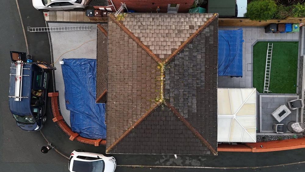 Aerial view of a house with a tiled roof, surrounded by a brick wall. Two cars are parked outside, a blue tarp covers parts of the driveway, and a green artificial lawn area is visible on the right.