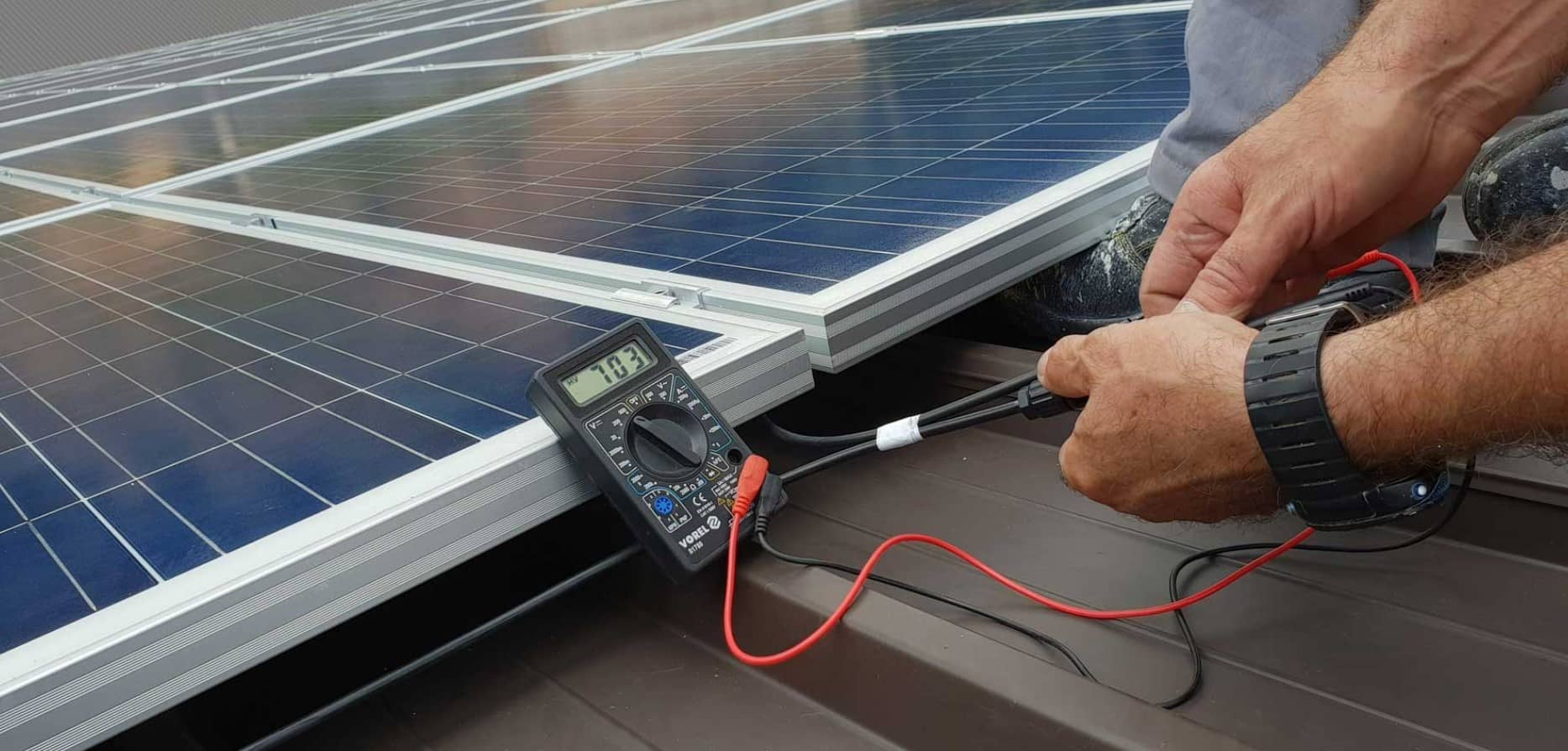 A person uses a digital multimeter to measure the voltage of solar panels installed on a rooftop. The multimeter display reads 17.3, and the person holds the test probes against the panel connections.