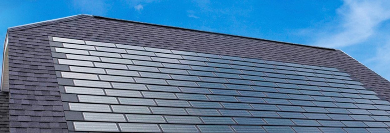 A sloped house roof is covered with sleek, rectangular solar shingles arranged in neat rows, blending in with traditional roof shingles under a clear blue sky.