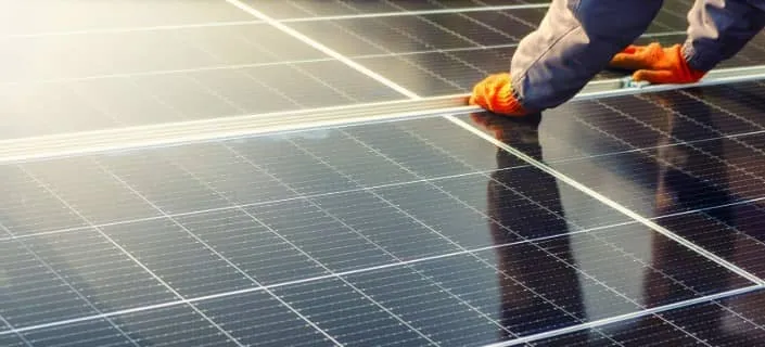 A person wearing orange gloves and a gray jacket installs or maintains solar panels, carefully positioning them outdoors in bright sunlight.
