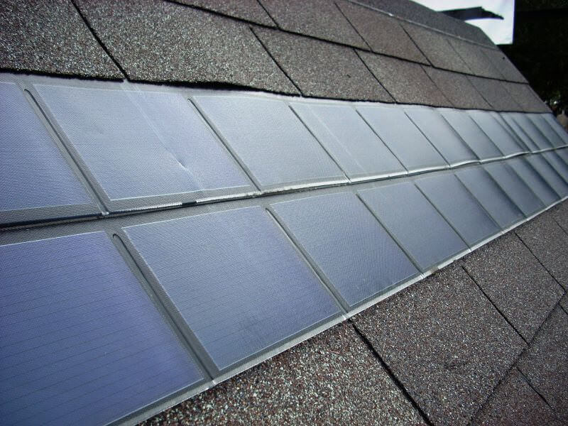 Solar panels integrated into the shingles of a sloped roof, showing a close-up view of the solar shingles aligned in rows among standard asphalt roofing tiles.