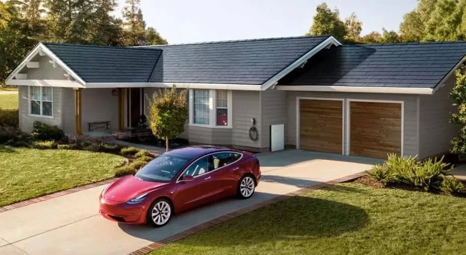 A modern single-story house with solar roof tiles, a double garage with wooden doors, and a red Tesla car parked in the driveway, surrounded by grass and trees on a sunny day.