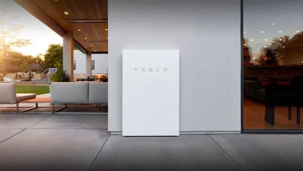 A white Tesla Powerwall battery is mounted on the exterior wall of a modern house, with a patio and outdoor seating area visible to the left and large windows to the right.