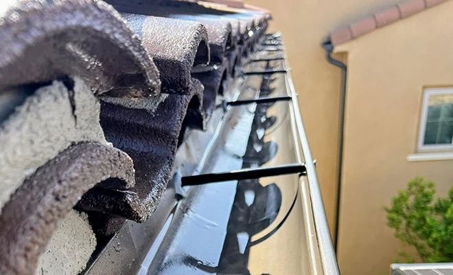 Close-up view of a house roof with curved tiles and an attached metal gutter system. The gutter is clean and has curved brackets for support. In the background, part of another house and some greenery are visible.