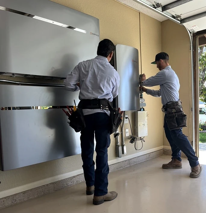 Two technicians in work uniforms and tool belts install or service electrical equipment mounted on a garage wall, with large metallic units and wiring visible. Daylight enters through the open garage door.
