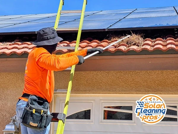 A worker in an orange shirt and hat stands on a ladder, cleaning debris from a house gutter near solar panels. A tool belt hangs at their waist. The "Solar Cleaning Pros" logo is in the bottom right corner.