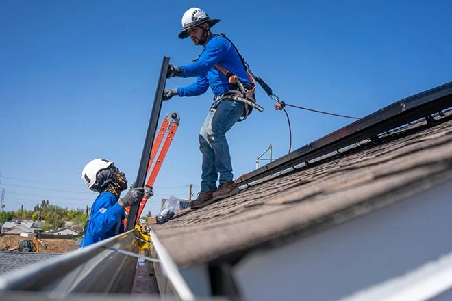 Two workers wearing helmets and safety harnesses install solar panels on a sloped roof under a clear blue sky, using tools and an orange ladder.